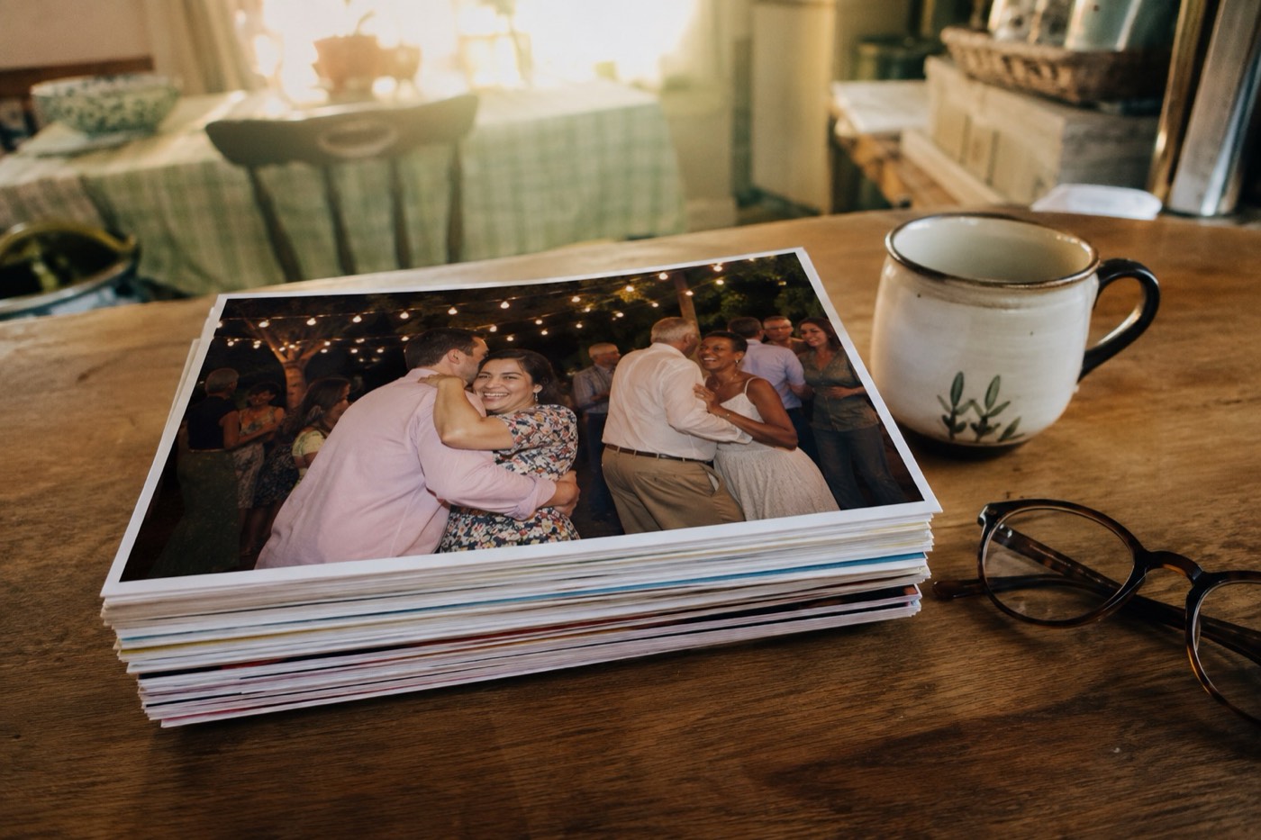 A thick stack of freshly printed party photographs on a kitchen table beside a mug and reading glasses