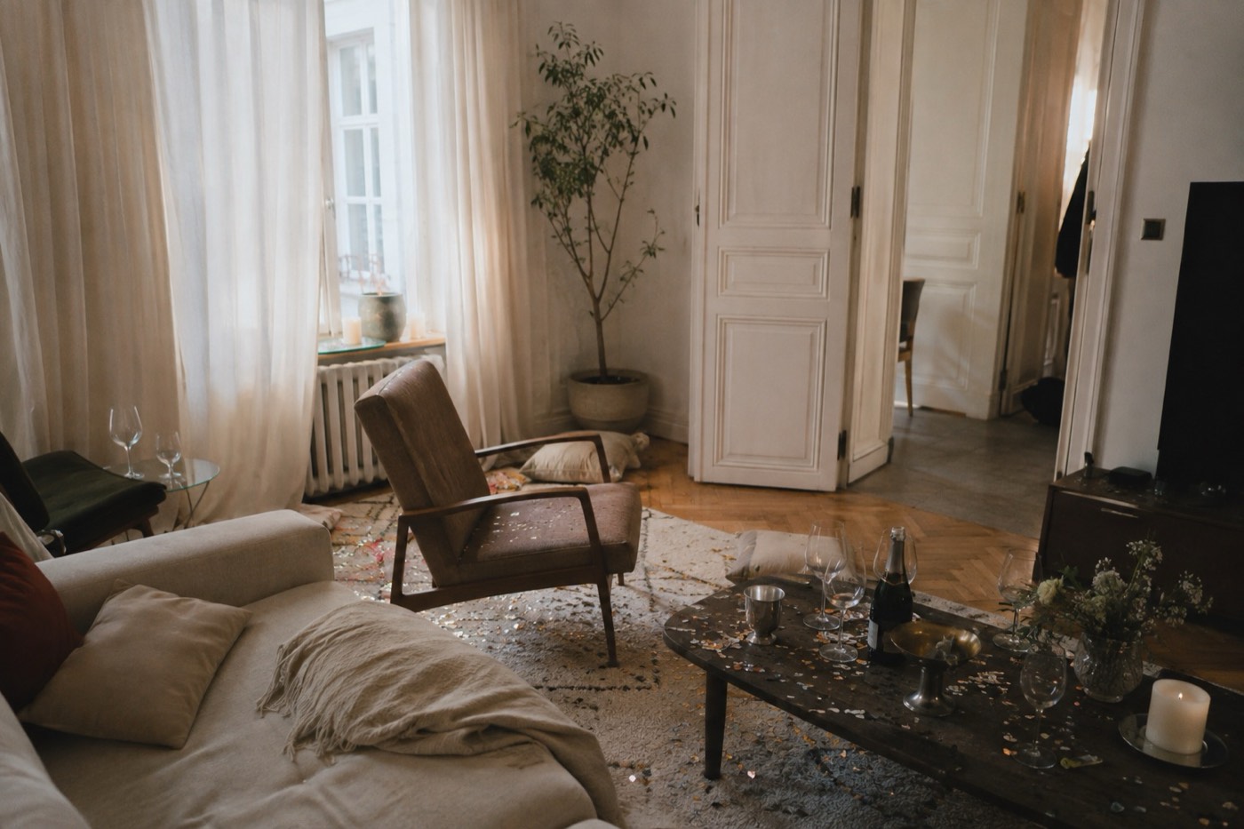 A living room the morning after a party, grey daylight, glasses and confetti still on the rug