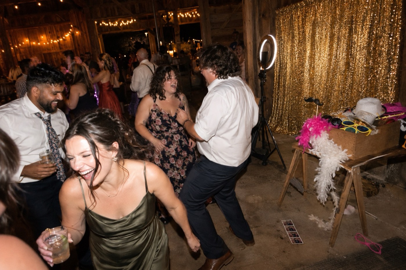 Guests dancing in a barn at a wedding late at night while the hired photo booth in the corner stands empty: a gold sequin backdrop, a ring light, a box of feather boas and party glasses, and a photo strip dropped on the floor