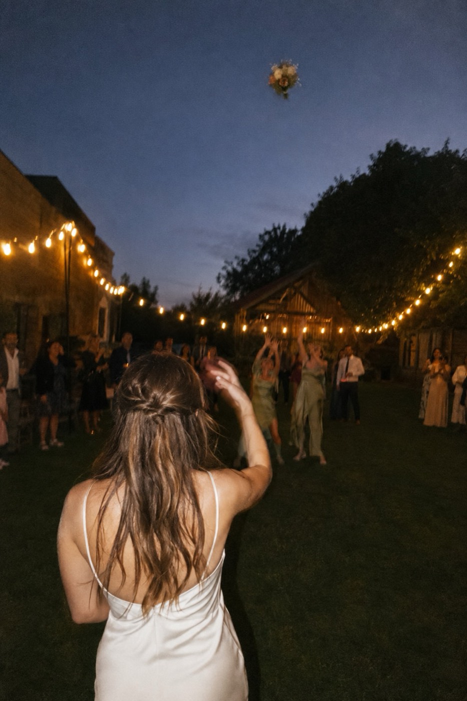 A bride seen from behind on the lawn at dusk, her bouquet in the air and a knot of guests reaching up for it