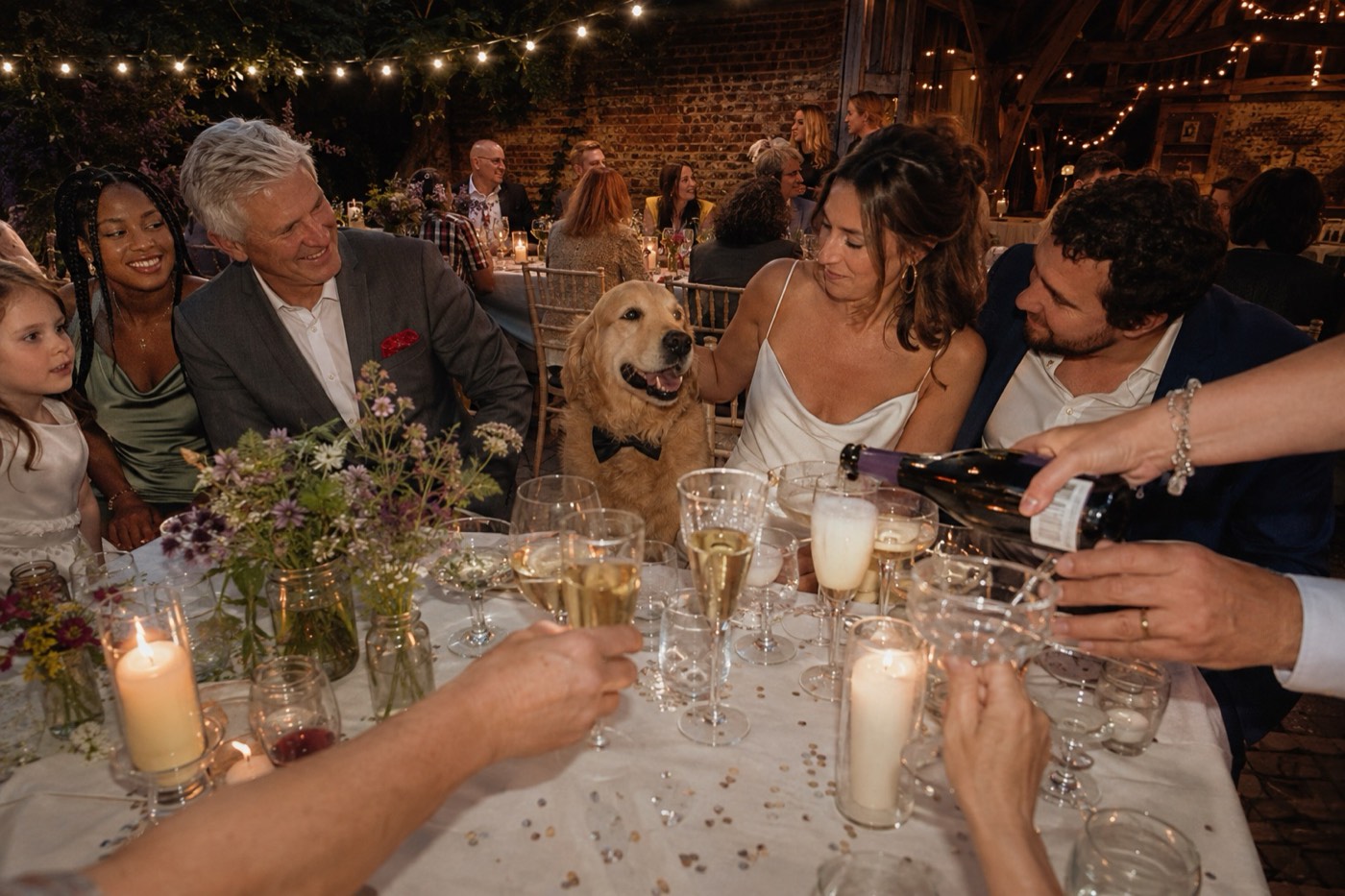 A crowd of mismatched glasses being filled at the end of the wedding table at night, hands reaching in to take them