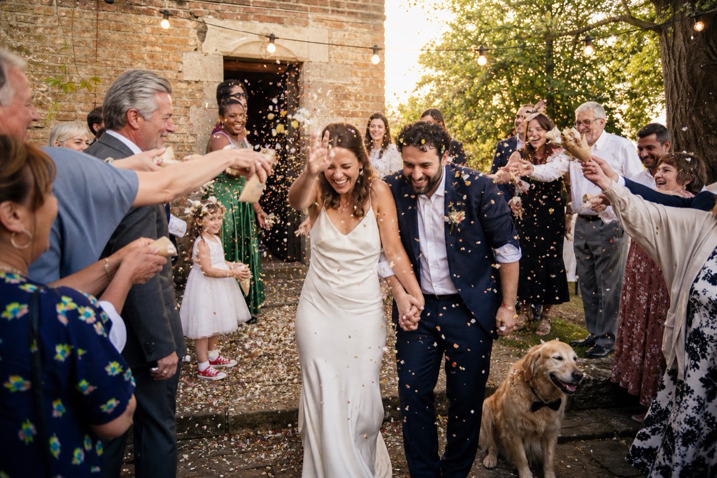 Guests throwing confetti over a bride and groom as they come out of the stone doorway of a country house in low afternoon sun