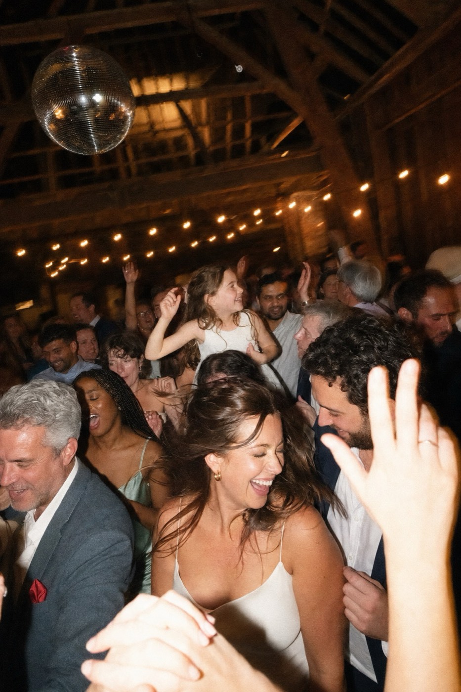 A packed barn dance floor late at night, flash-lit and smeared by the slow shutter, the bride barefoot in the middle of it