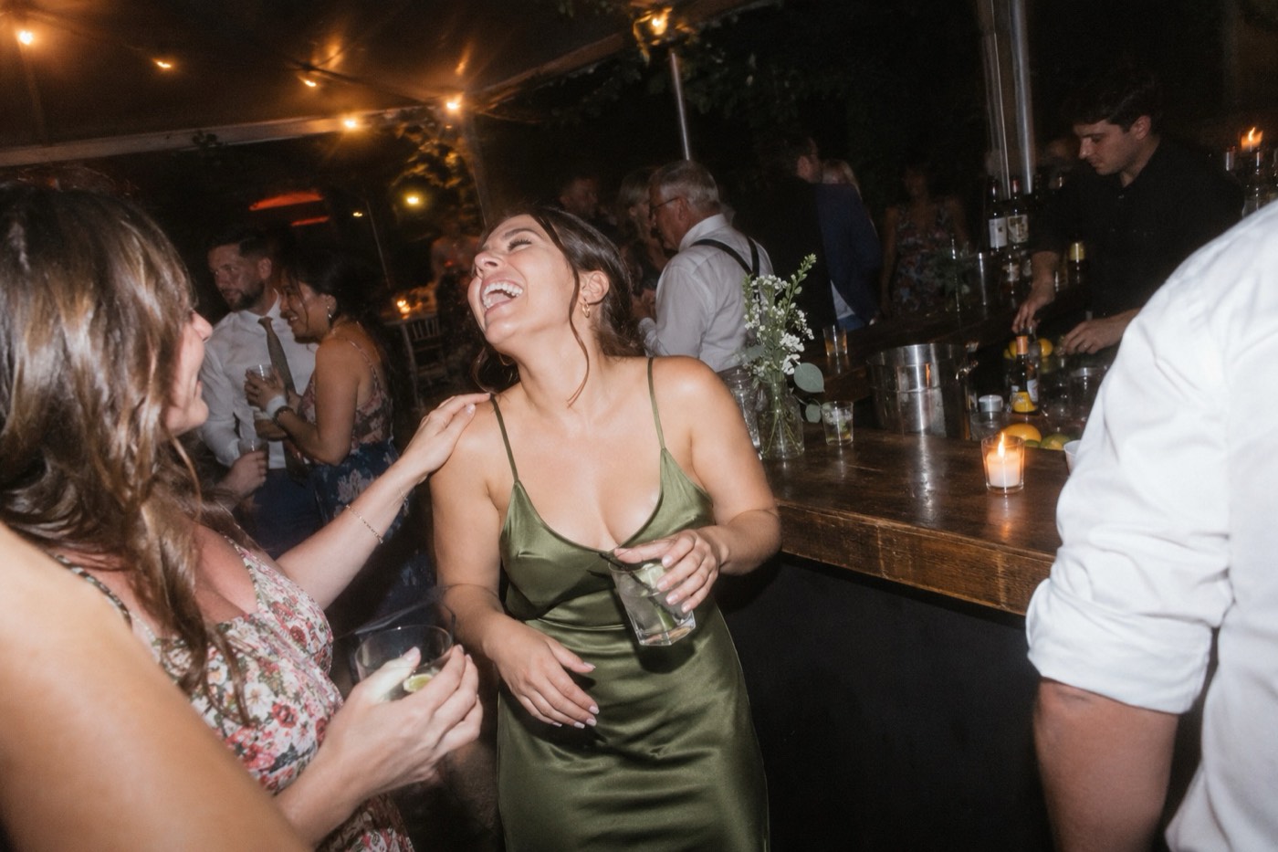 A wedding guest in a green satin dress laughing with her head thrown back at the bar late at night, lit by a hard flash, a friend’s hand on her shoulder