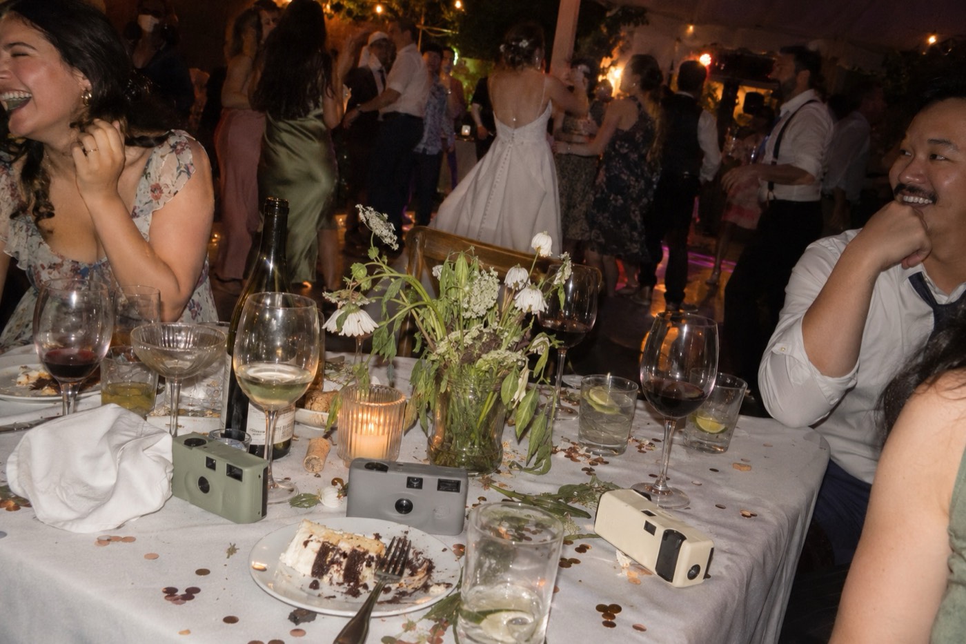 A wedding table at the end of the night covered in confetti, half-empty glasses and cake, with three plain plastic disposable cameras left among the glasses while the bride dances beyond