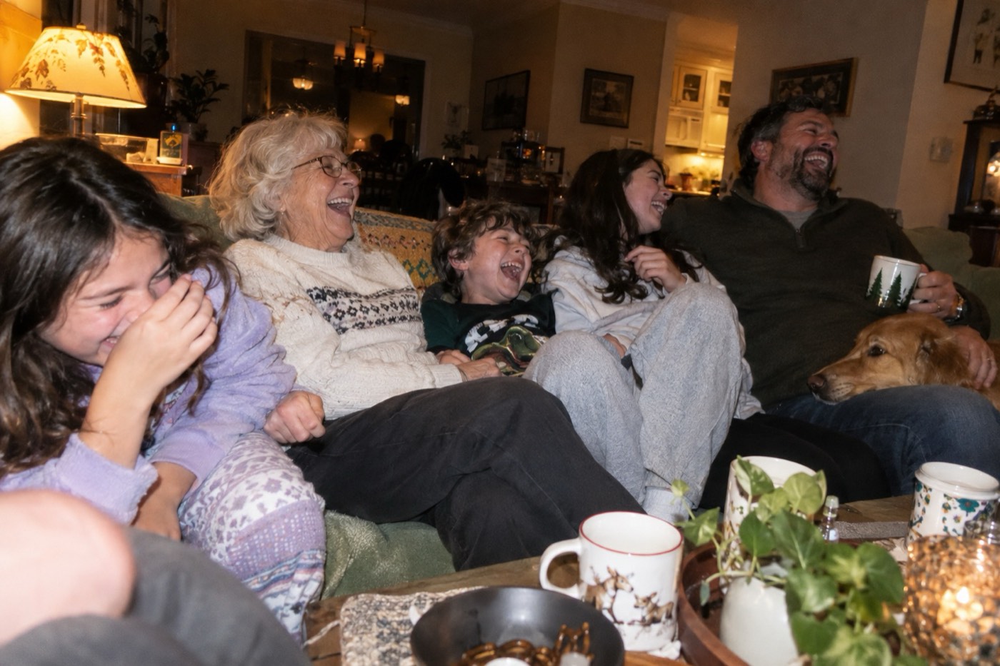 A grandmother squeezed onto a sofa with her grandchildren and grown-up son, all of them helpless with laughter, a dog resting its head on a knee