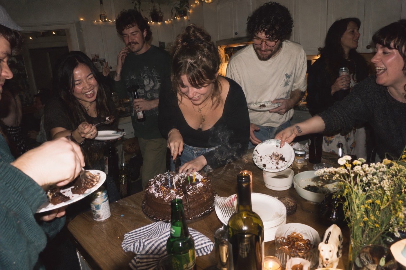 Friends crowded round a kitchen table at a birthday party at night, one cutting a chocolate cake as the candles smoke, others reaching in with paper plates