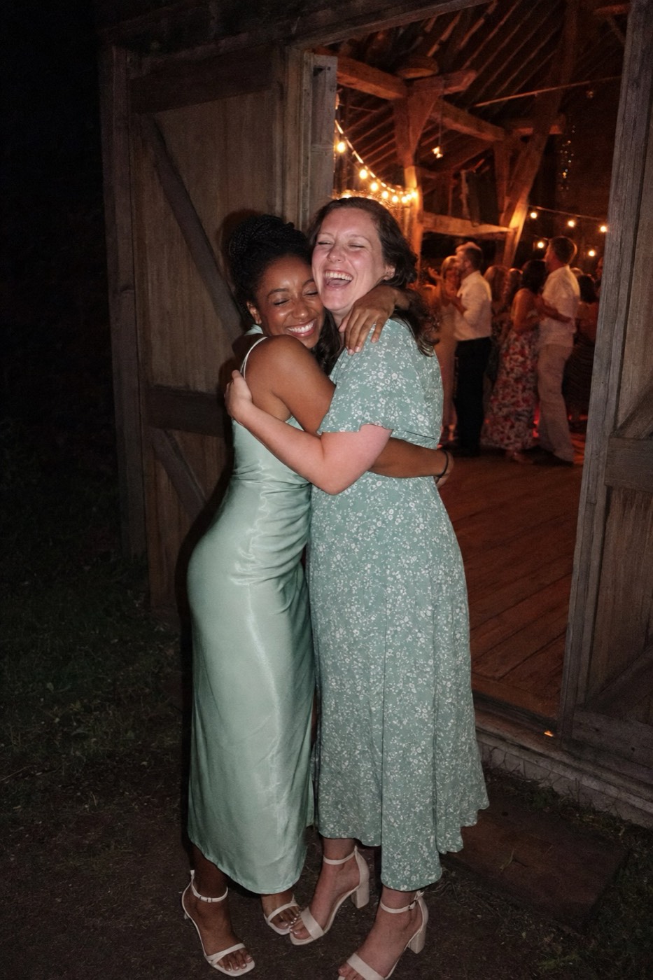 Two women guests hugging hard in the open doorway of the barn, the lit dance floor blurred behind them