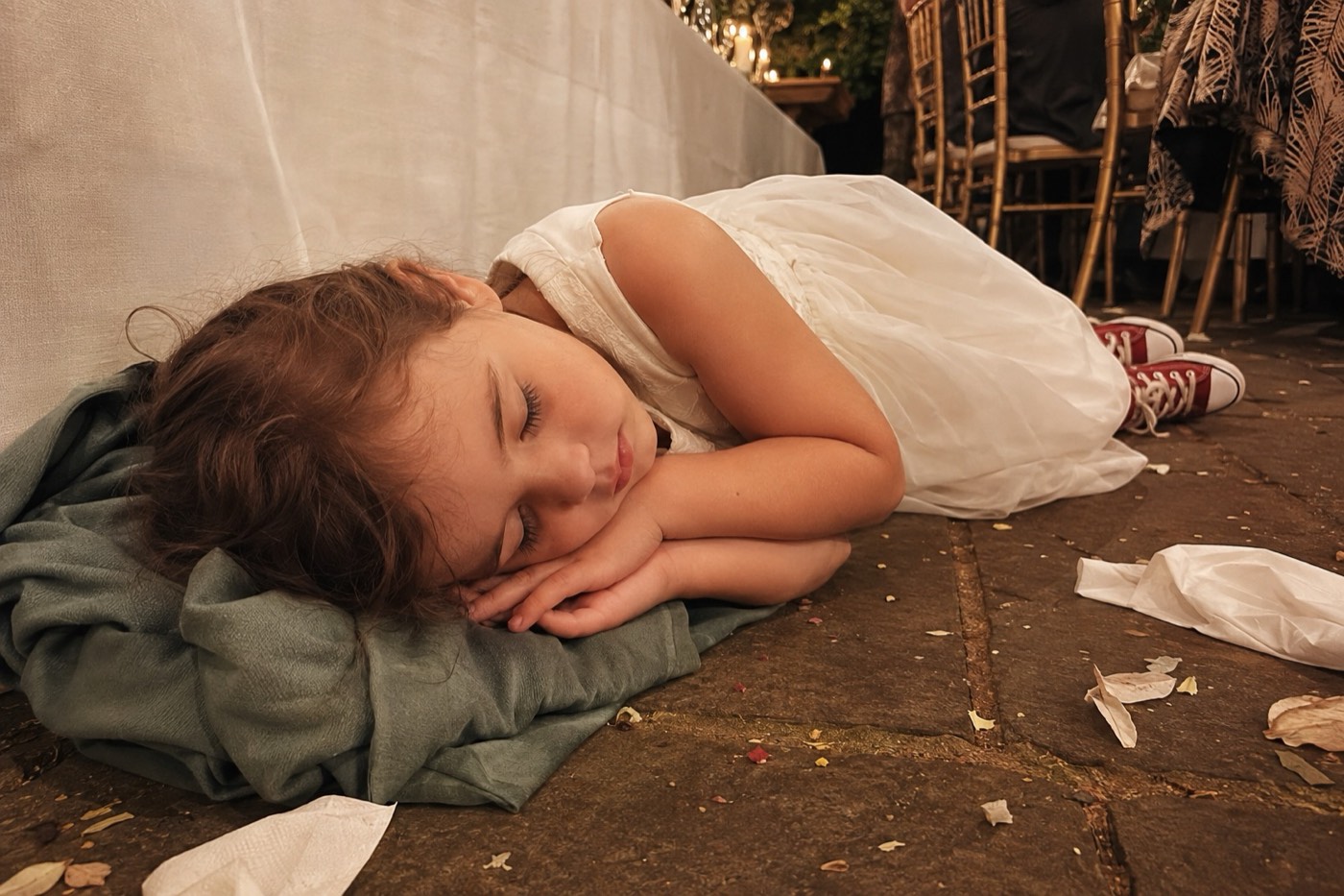 A flower girl in a white dress and red trainers fast asleep on a folded coat beside the wedding table, among fallen confetti and kicked-off shoes