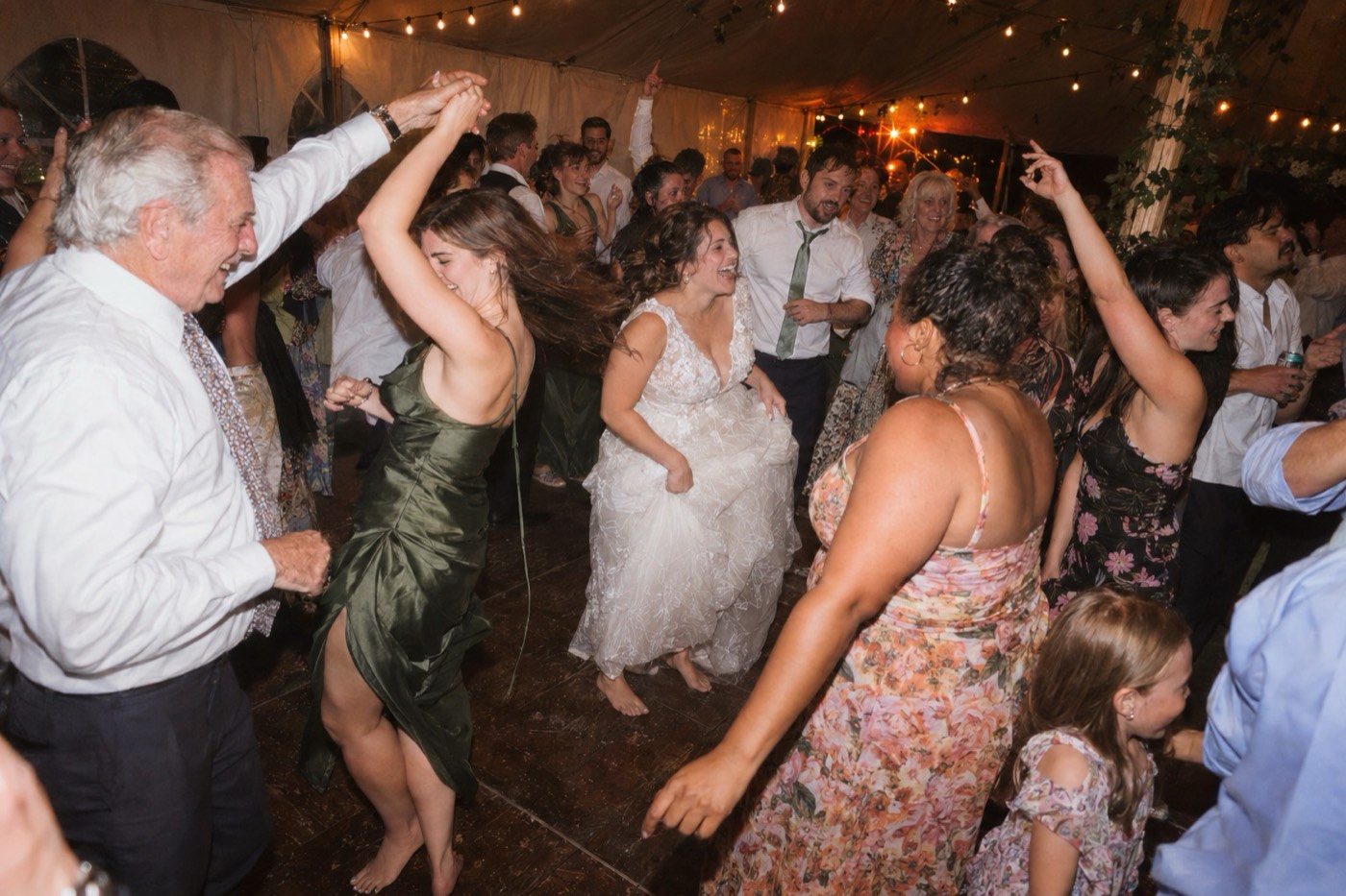 A packed dance floor in a wedding marquee at night, the barefoot bride laughing in the middle and an older guest twirling a woman in a green dress