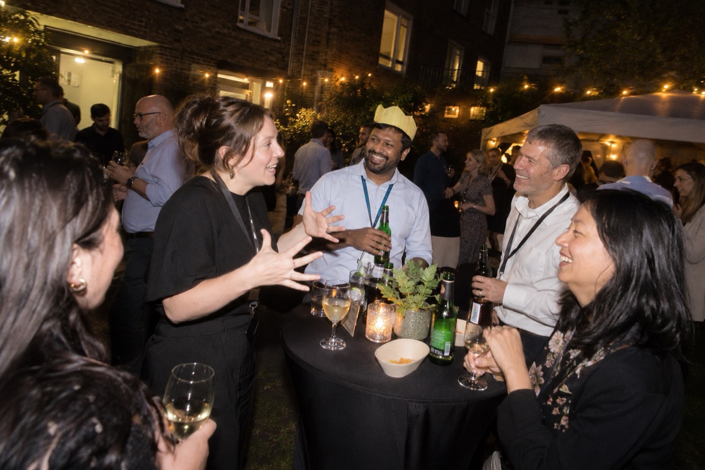 Colleagues with lanyards laughing round a high table at a company summer party in an office courtyard under string lights, one in a paper crown