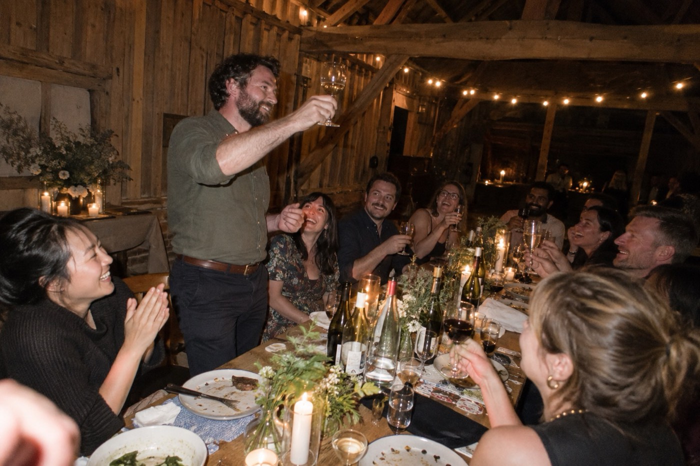 A team dinner at a long candlelit table in a wooden barn, one colleague standing to give a toast while the others raise their glasses and cheer