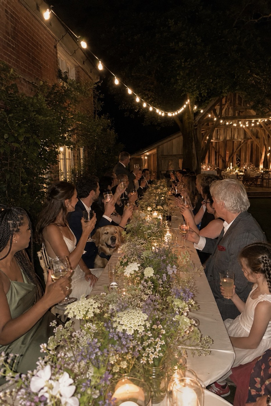 Looking down the length of the candlelit wedding table at night, guests either side turned in with their glasses raised