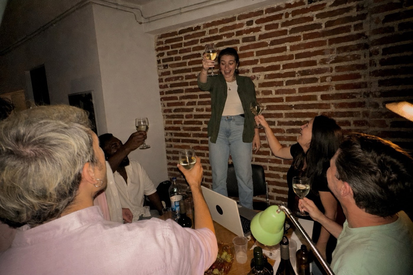 A small team crowded round a table in a back room, one of them standing on a chair mid-toast