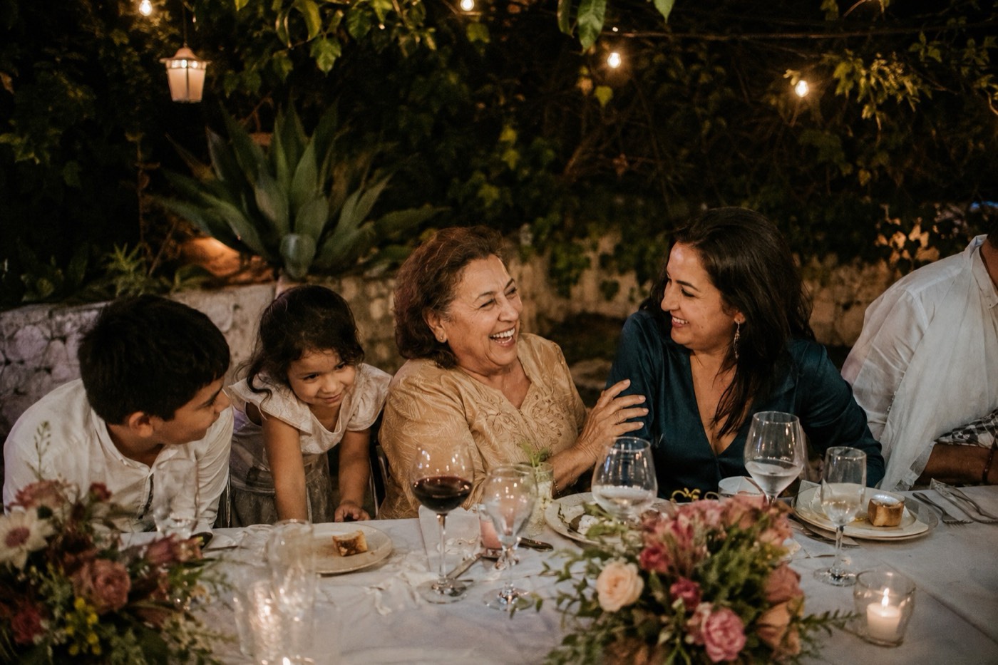 Three generations at a long garden table, a grandmother laughing with two children leaning in over the cloth