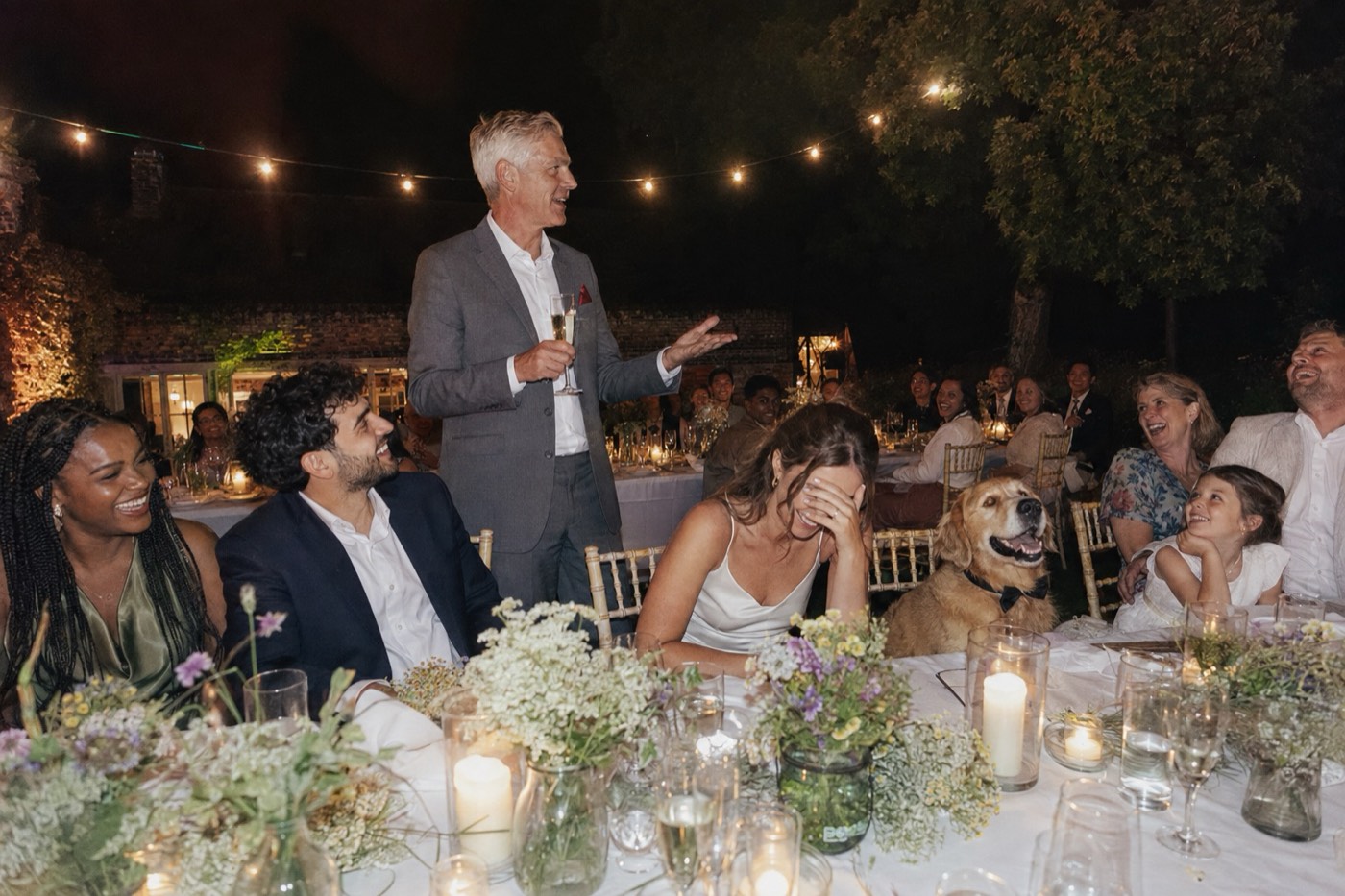 The best man on his feet mid-speech at the table, one hand out, the guests either side of him laughing
